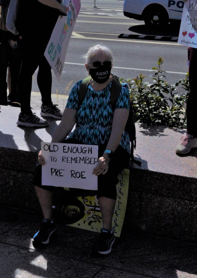 An elderly protester sits holding a sign reading “old enough to remember pre-Roe,” referring to the Supreme Court case Roe v. Wade. 