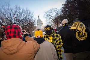 A photographer frames a shot of the Capitol as Proud Boys look on.